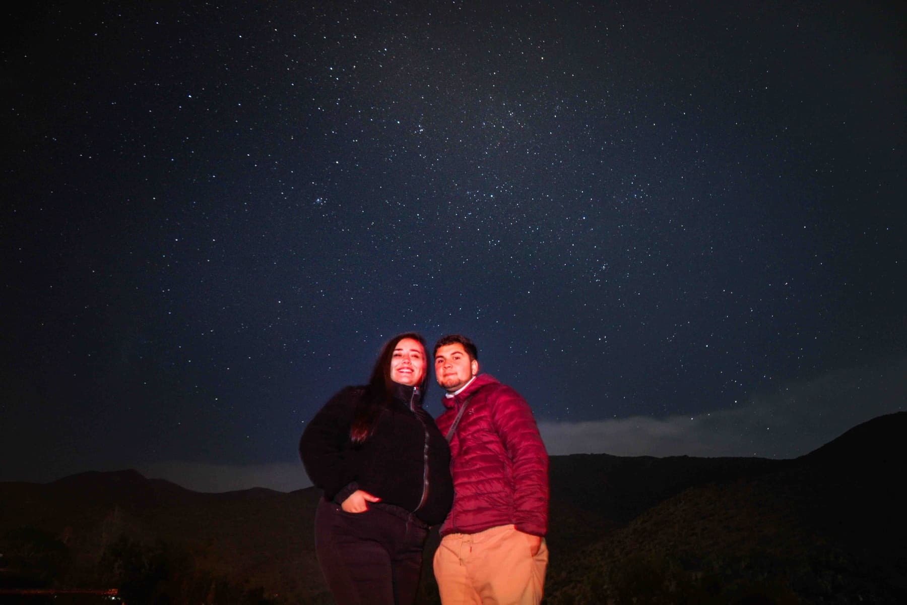 Una pareja sonríe bajo un cielo lleno de estrellas, con los cerros del valle al fondo.