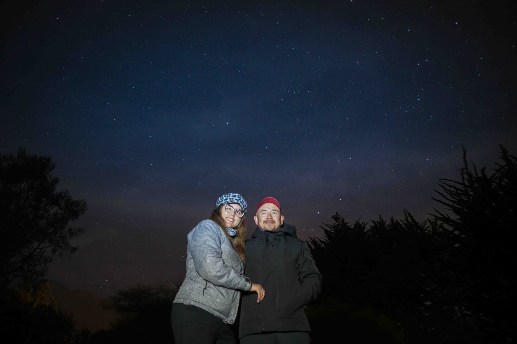 Una pareja abrigada posa bajo las estrellas, entre la silueta de los árboles.