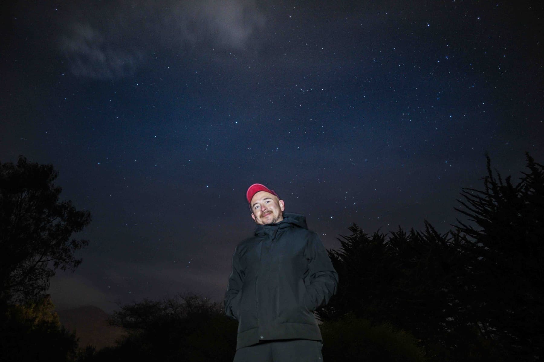 Un visitante con gorro rojo, de pie bajo el cielo estrellado entre los árboles.